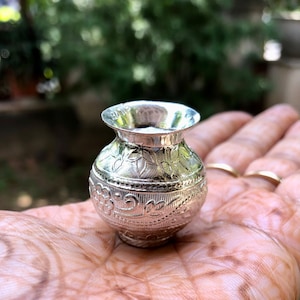 Może przedstawiać: Mały, srebrny, ozdobny wazon z misternymi wzorami kwiatowymi. Wazon ma około 5 cm wysokości i okrągły korpus ze lekko rozszerzonym wierzchołkiem.