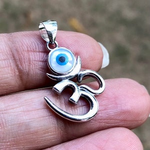 May include: Silver pendant featuring an Om symbol with a blue evil eye charm. The eye is made of mother of pearl and has a black pupil.