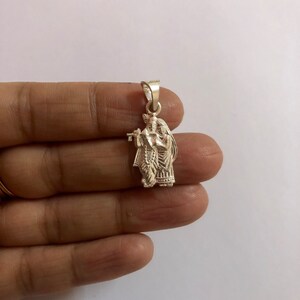 Op de afbeelding: Een zilveren hanger met een gedetailleerde afbeelding van Krishna en Radha, een hindoe&iuml;stisch godenpaar, waarbij Krishna een fluit vasthoudt.