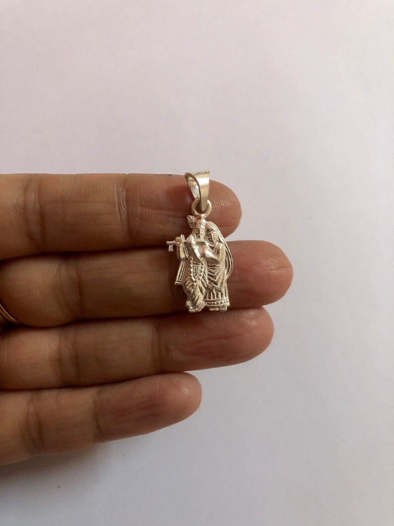 Op de afbeelding: Een zilveren hanger met een gedetailleerde afbeelding van Krishna en Radha, een hindoe&iuml;stisch godenpaar, waarbij Krishna een fluit vasthoudt.