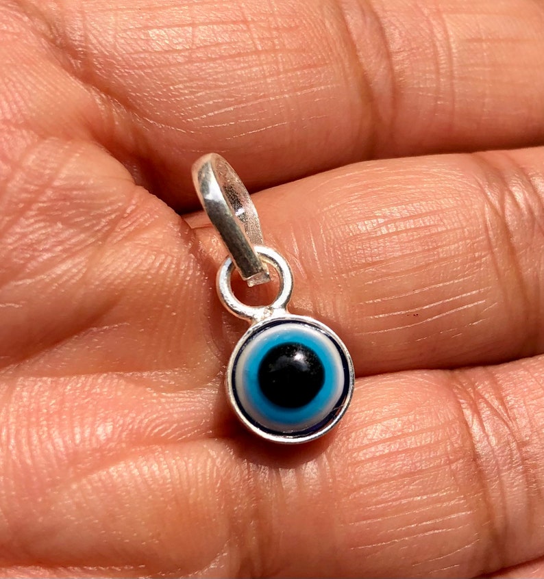 Puede incluir: Un amuleto de plata con un dise&ntilde;o de ojo turco azul. El amuleto es redondo y tiene un bucle de plata en la parte superior para sujetarlo a un collar o pulsera.