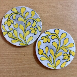 May include: Two round ceramic coasters with a white background and a yellow floral design. The flowers are outlined in blue.