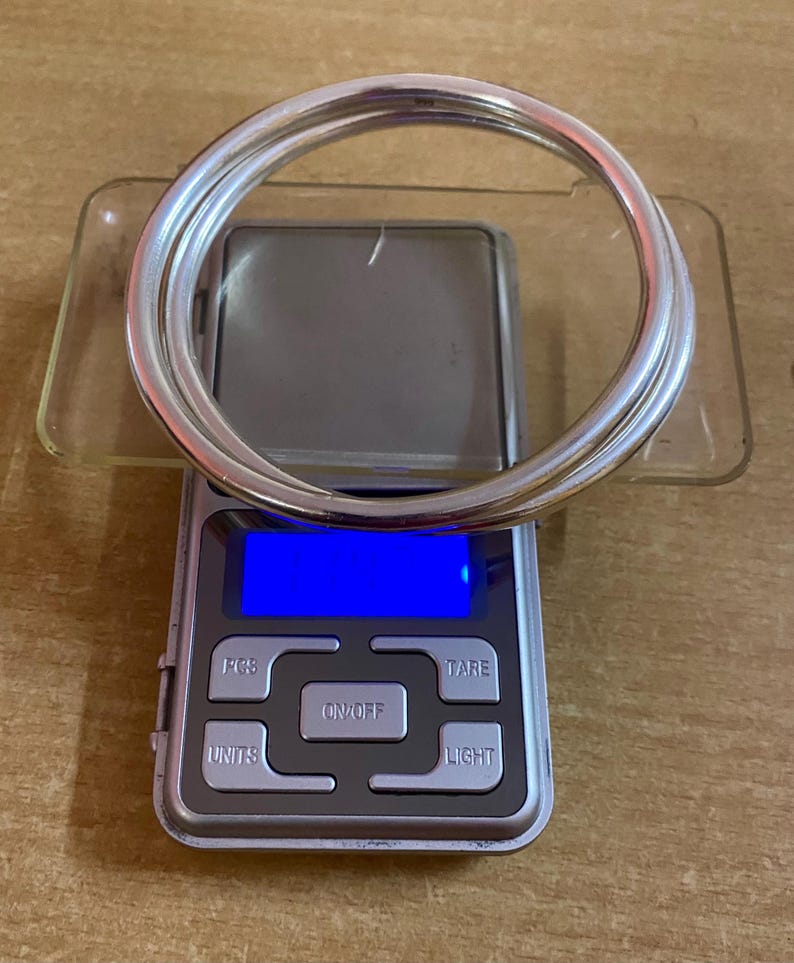May include: Two silver bangles on a digital scale with a blue display. The scale has buttons labeled "PCS", "TARE", "ON/OFF", "UNITS", and "LIGHT".