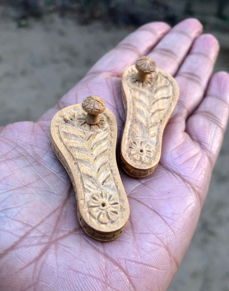 Pu&ograve; includere: Due piccole sandali di legno intagliate con un motivo floreale. Le sandali sono di colore marrone chiaro e hanno un piccolo pomo nella parte superiore.
