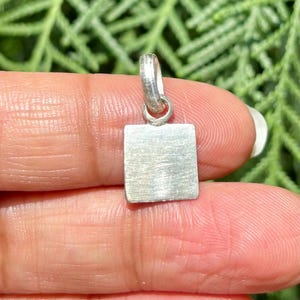 以下が含まれることがあります： 正方形の形をした小さな銀色のペンダント。チェーン用のループが付いています。ペンダントはつや消し金属仕上げで、少しざらざらした外観です。背景はぼやけた緑の葉です。