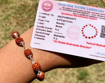 May include: A brown Rudraksha bracelet with five beads, made of natural Rudraksha seeds, is shown on a person's wrist. The bracelet is 6.670 grams and is certified by the ISKON Gem Testing Laboratory. The certificate states that the bracelet is a "(5) Mukhi Rudraksha Bracelet".