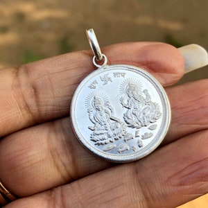 Puede incluir: Pendiente de plata con una representación de Lakshmi y Ganesha, dos deidades hindúes, con la palabra sánscrita "Laxmi" inscrita alrededor del borde.