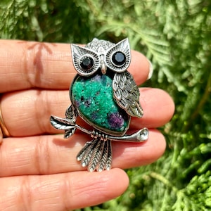 Könnte beinhalten: Silberne Eulenbrosche mit einem Körper aus grünem und lila Edelstein. Die Eule hat schwarze Edelsteinaugen und sitzt auf einem Ast. Die Brosche ist mit Feder- und Blattmotiven versehen, ein einzigartiges Schmuckstück.