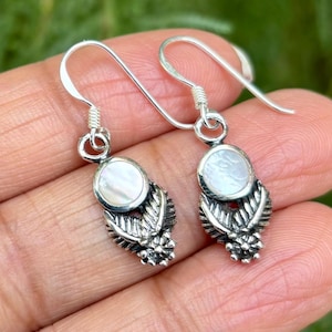 May include: A pair of silver earrings with a white mother of pearl stone and a detailed silver design. The earrings have a hook closure.