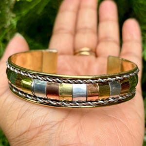 May include: A gold-coloured cuff bracelet with a decorative band featuring alternating rectangular inlays of silver, copper, and gold. The bracelet has a braided rope design along the edges and is open at the bottom.