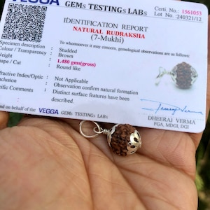 May include: A brown Rudraksha bead with a silver pendant, weighing 1.480 grams. The bead is studded and has a round shape. The image includes a certificate of authenticity from VEGGA GEMS TESTINGS LABS, stating that the Rudraksha is natural and has 7 Mukhi.