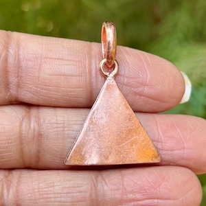 May include: A copper triangle pendant with a smooth, slightly textured surface. The pendant has a loop at the top for attaching to a chain.