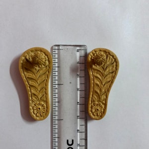 Pu&ograve; includere: Due sandali in legno intagliati con intricati motivi floreali. I sandali sono lunghi circa 5 cm e sono realizzati in legno marrone chiaro.