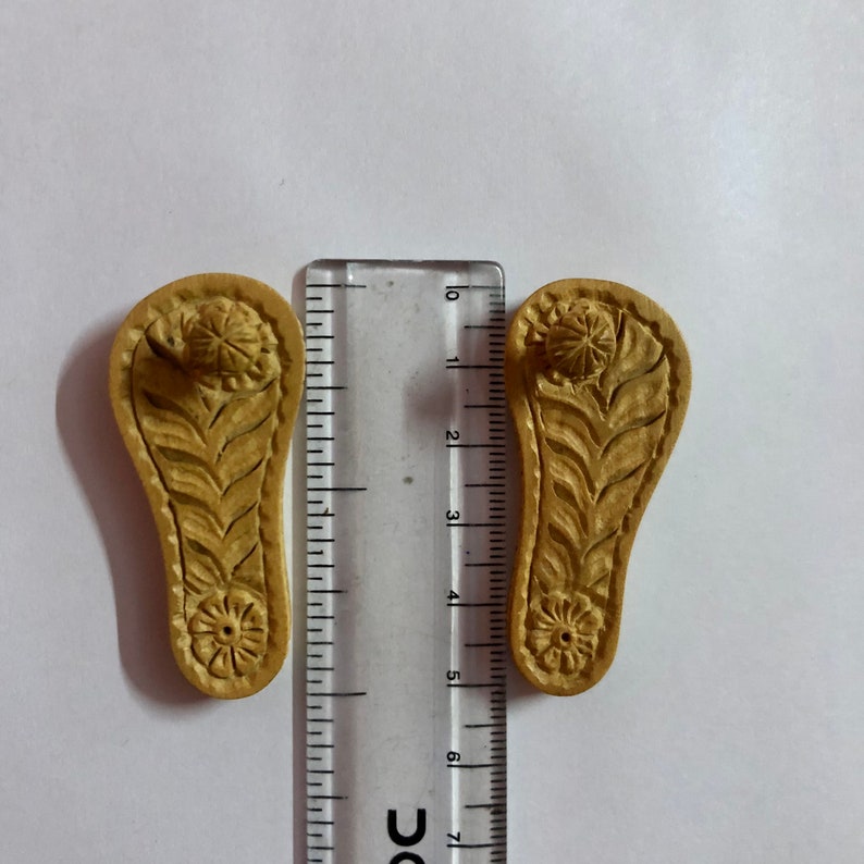 Pu&ograve; includere: Due sandali in legno intagliati con intricati motivi floreali. I sandali sono lunghi circa 5 cm e sono realizzati in legno marrone chiaro.