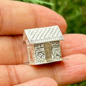 Puede incluir: Un amuleto de plata con forma de casita pequeña con un techo detallado y una puerta abierta. El amuleto se sostiene en una mano.
