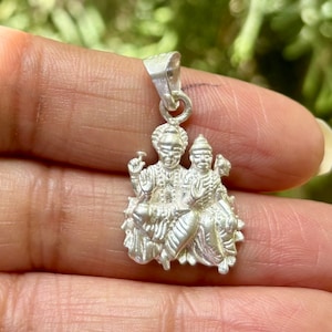 May include: A silver pendant featuring two figures in detailed relief. The pendant has a loop for a chain and is held in a hand against a blurred green background. The figures are seated and adorned with intricate clothing and headdresses.