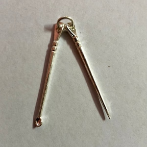 May include: A silver charm in the shape of a pair of crossed tweezers. The charm has a loop at the top for attaching to a necklace or bracelet.
