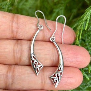May include: A pair of silver dangle earrings with a delicate, intricate design. The earrings are shaped like a curved line with a floral pattern at the bottom.