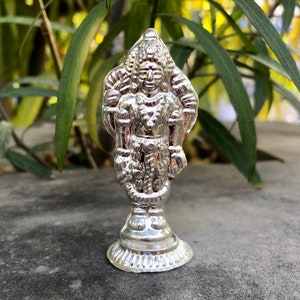 May include: A silver figurine of a Hindu deity standing on a pedestal. The deity is wearing a crown and elaborate jewelry. The figurine is detailed and intricate.