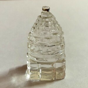 Puede incluir: Una pirámide de cristal transparente con forma de templo, con una base cuadrada y una punta puntiaguda. El cristal es transparente y tiene un acabado liso y pulido.