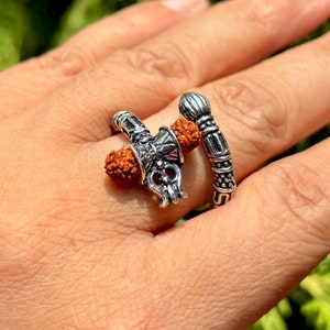 Peut inclure: Une bague en argent avec un motif de trident et de bâton, tous deux ornés de perles rouges. La bague est portée sur un doigt.