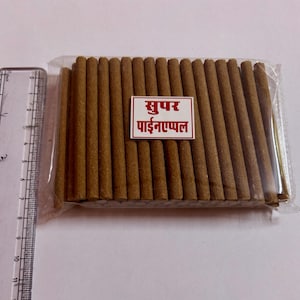 May include: A package of brown incense sticks, tightly packed and wrapped in clear plastic. A small rectangular label with red and white text is visible. A ruler is placed next to the package, showing measurements in centimetres.