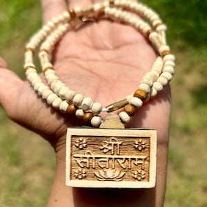 May include: A white and brown wooden beaded necklace with a wooden pendant featuring the Sanskrit text "Shree Sita Ram" in a carved design.