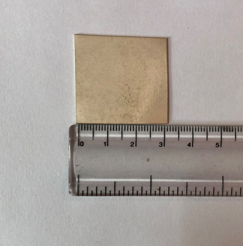 May include: A gold-colored metal sheet measuring approximately 3 inches by 3 inches. The sheet is placed on top of a ruler for size reference.