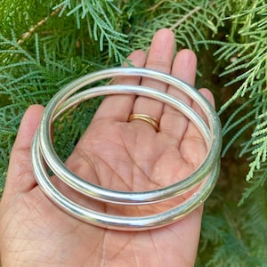 May include: Two silver bangles, one slightly larger than the other, are stacked on top of each other. The bangles are smooth and shiny, and they are held in a person's hand.