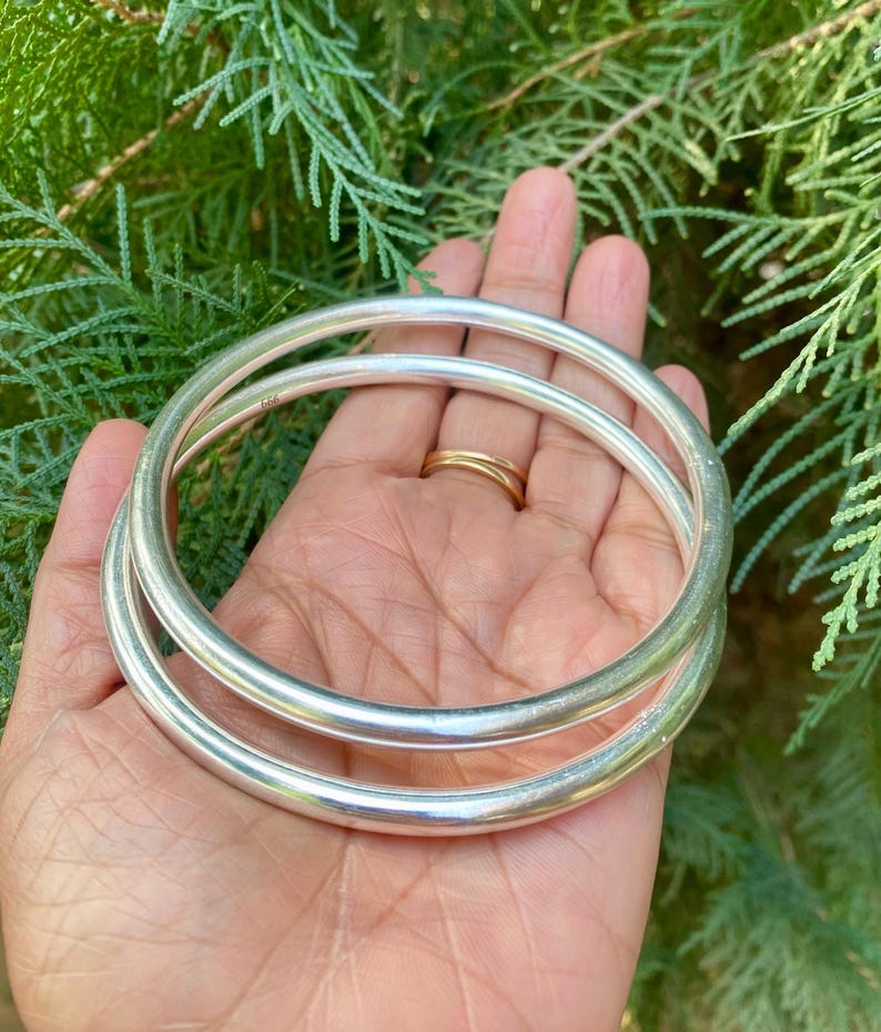 May include: Two silver bangles, one slightly larger than the other, are stacked on top of each other. The bangles are smooth and shiny, and they are held in a person's hand.
