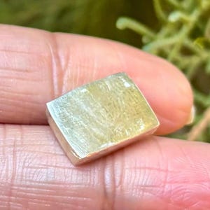 May include: A small, rectangular, light green gemstone with a polished surface. The gemstone has a subtle, natural pattern and is held between fingers, showcasing its size and colour.