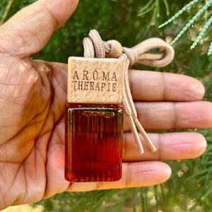 Op de afbeelding: Een bruine houten autoluchtverfrisser met de tekst "Aroma Therapie" erop. De luchtverfrisser is bevestigd aan een beige touwtje en hangt aan een hand.