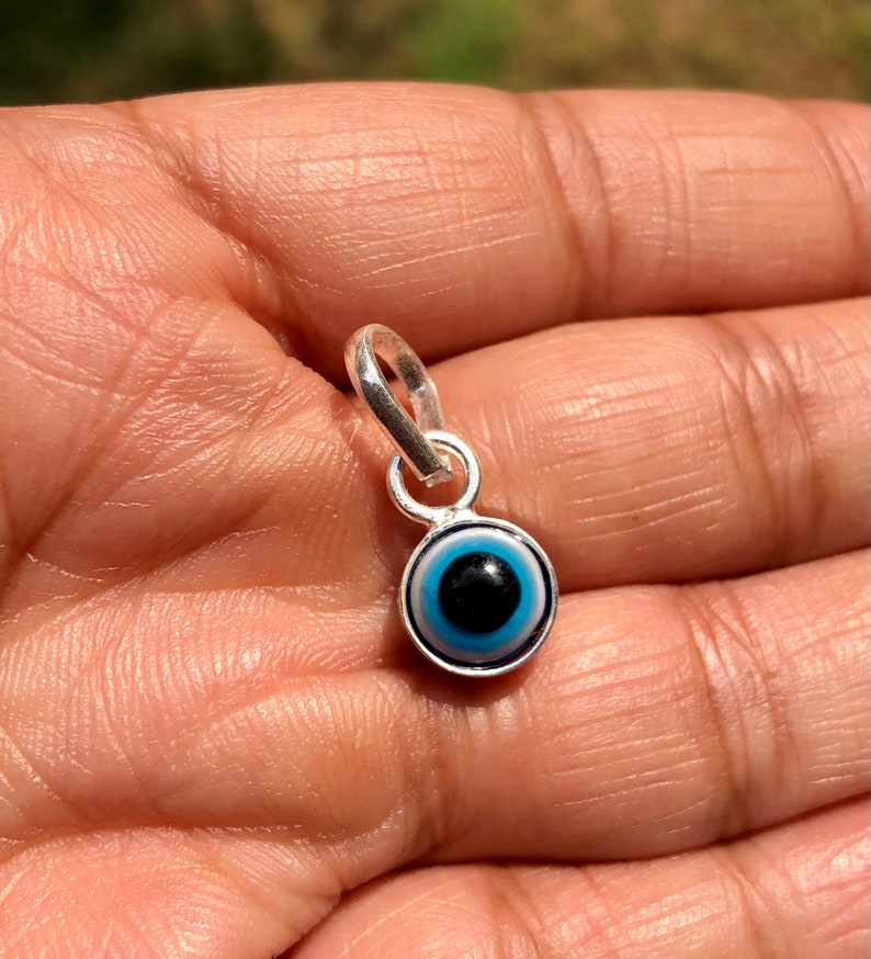 Puede incluir: Un amuleto de plata con un dise&ntilde;o de ojo azul malvado. El amuleto tiene un bucle en la parte superior para sujetarlo a una pulsera o collar.