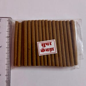 May include: A transparent packet containing numerous brown incense sticks. The sticks are cylindrical and tightly packed. A red and white label with text is attached to the packet. A ruler is on the left.