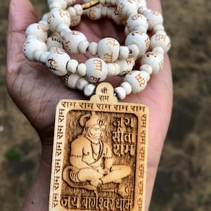 May include: A wooden mala necklace with white beads and a wooden pendant featuring a carved image of Hanuman, a Hindu deity. The pendant has the text "Shri Ram" and "Jai Sita Ram" in Hindi.