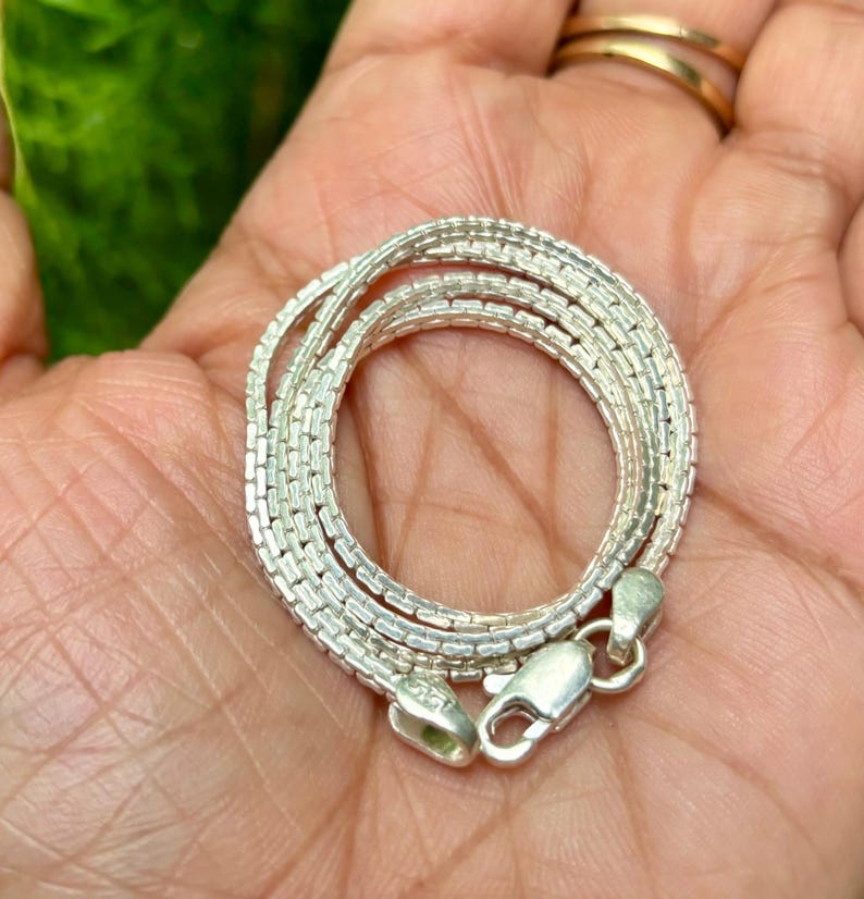 May include: A silver chain necklace with a box chain design. The chain is coiled in a hand.