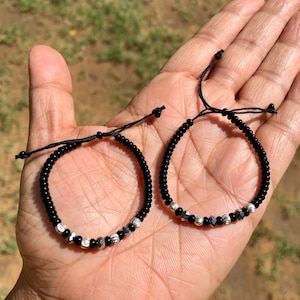 Könnte beinhalten: Zwei schwarze Perlenarmbänder mit silbernen Akzenten. Die Armbänder sind mit schwarzem Band gebunden und haben ein schlichtes Design.