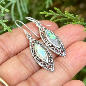 May include: A pair of silver dangle earrings with a pearlescent abalone shell inlay. The earrings have a delicate filigree design.