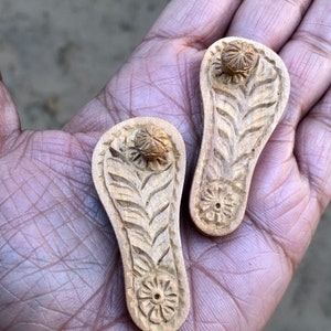 Pu&ograve; includere: Due porta incenso in legno intagliati a forma di sandali con intricati motivi floreali. I porta incenso sono di colore marrone chiaro e hanno un piccolo foro al centro della parte inferiore.