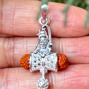 Può includere: Ciondolo in argento con una rappresentazione della divinità indù Shiva, che tiene un trishul e un damaru, con perline di rudraksha su entrambi i lati. Il ciondolo ha una piccola sfera d'argento appesa alla parte inferiore.