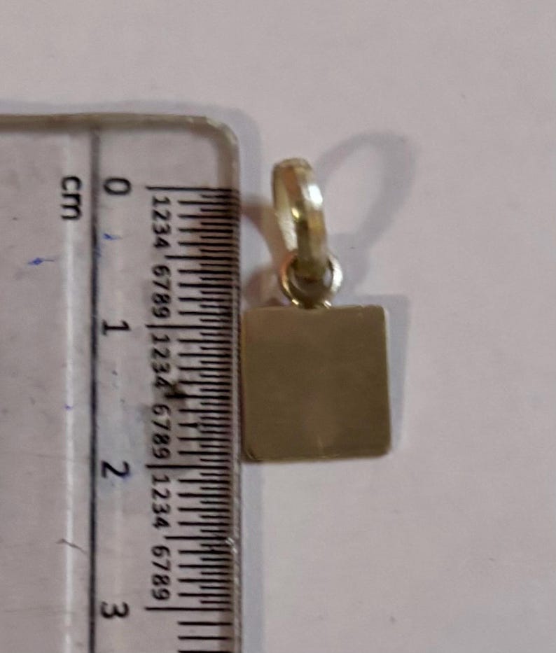 以下が含まれることがあります： チェーン用のループが上部にある、小さな金色の正方形のペンダント。 ペンダントは定規の隣にあり、高さは約2cmです。 ペンダントはおそらく金属製で、表面は滑らかです。