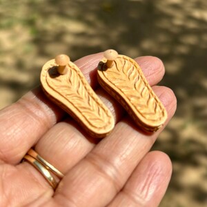 May include: A pair of wooden earrings shaped like sandals with a carved design. The earrings are a light brown color and have a small post on the back for attaching to the ear.
