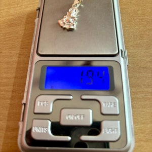 May include: A silver pendant shaped like a person sitting down, weighing 1.94 grams on a digital scale.
