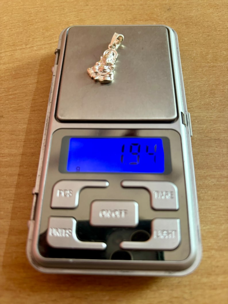 May include: A silver pendant shaped like a person sitting down, weighing 1.94 grams on a digital scale.