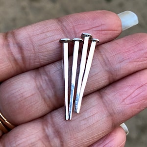 Puede incluir: Cinco tachuelas de metal plateado con cabeza plana, dispuestas en forma de abanico, sostenidas en una mano.