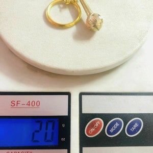 May include: A gold-coloured keychain with a small, round, textured charm. The keychain is on a digital scale that reads 20 grams.