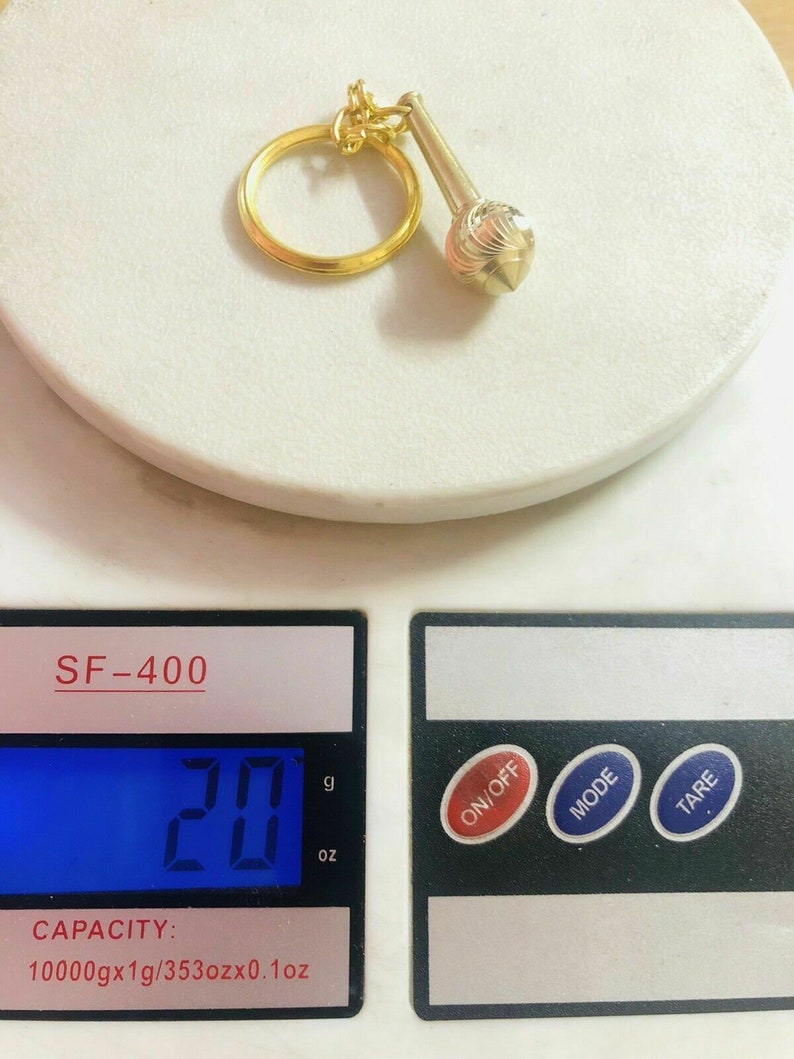 May include: A gold-coloured keychain with a small, round, textured charm. The keychain is on a digital scale that reads 20 grams.