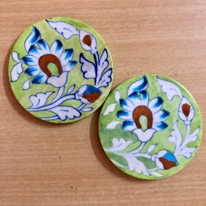 May include: Two round ceramic coasters with a green background and white floral designs. The flowers have blue and brown accents.