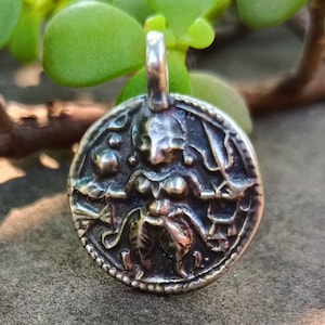 May include: Silver pendant featuring a detailed depiction of a Hindu deity, possibly Durga, with multiple arms and a fierce expression. The pendant is circular and has a textured border.