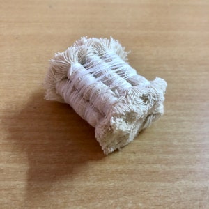 Op de afbeelding: Close-up van een bundel crèmekleurig katoenen touw, strak gewikkeld en vastgezet. Het touw heeft een getextureerde, gerafelde uitstraling aan de uiteinden, wat wijst op een handgemaakte of natuurlijke esthetiek. De bundel ligt op een lichtbruin houten oppervlak.